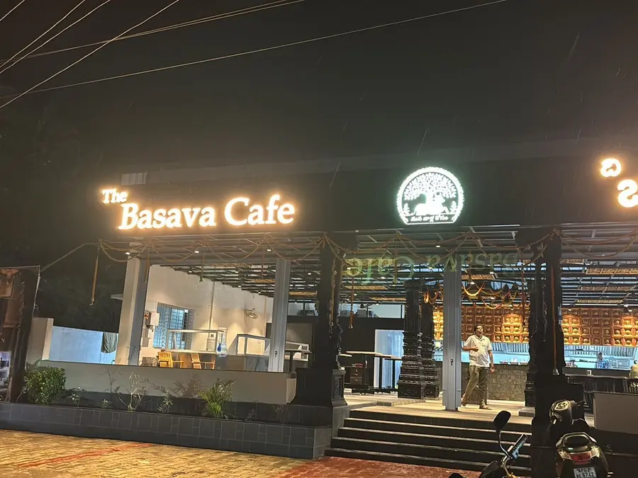 The Basava Cafe at dusk: the name lit in white above carved black pillars, a gold Nandi roundel glowing, marigold garlands strung across the front.
